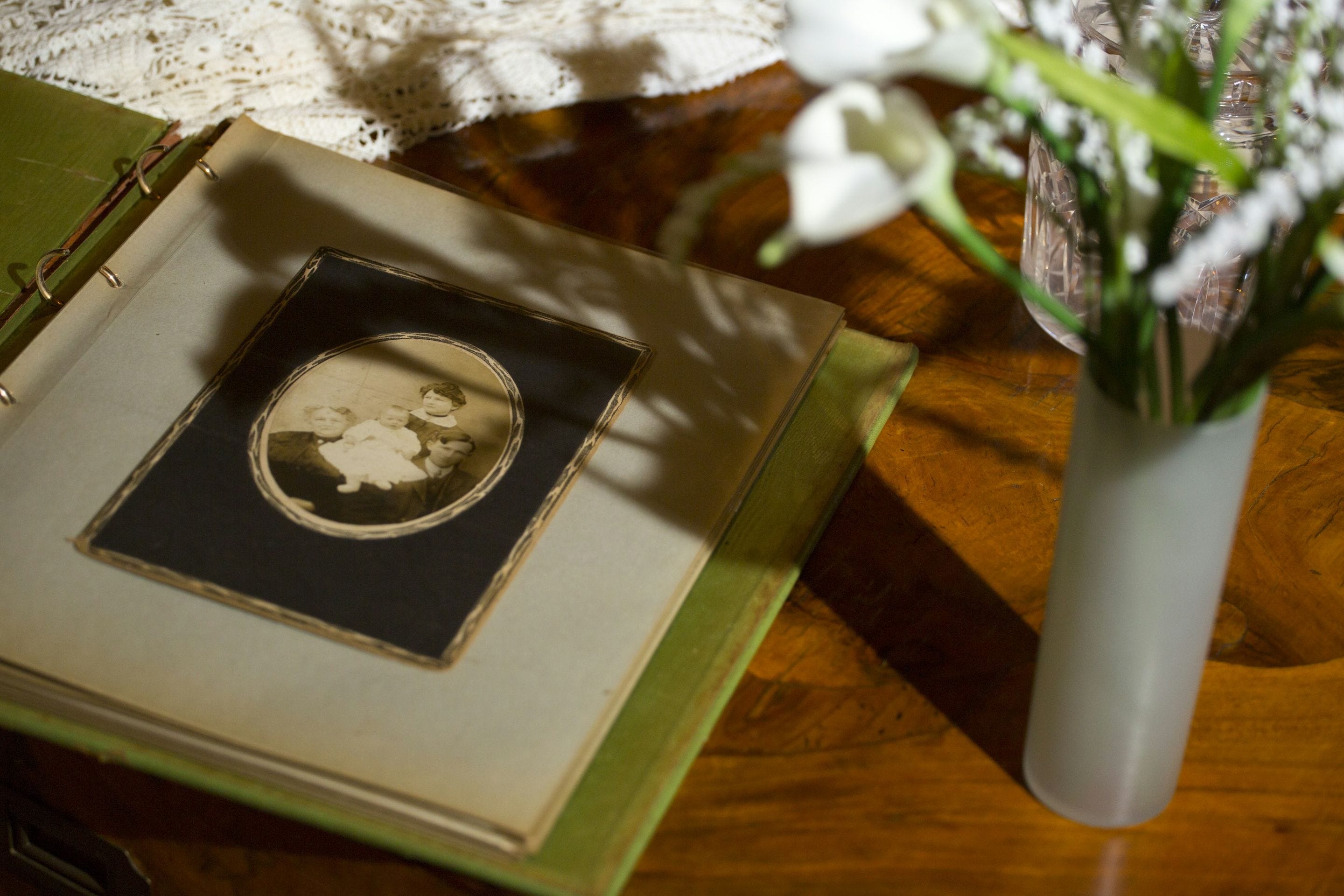 An open family album with an old portrait, beside white lilies on a lace-covered table in afternoon light